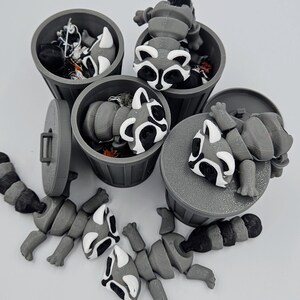 May include: A collection of gray 3D-printed raccoon figurines, some inside miniature trash cans and others disassembled. The raccoons have black and white markings. The trash cans are gray with lids and handles.