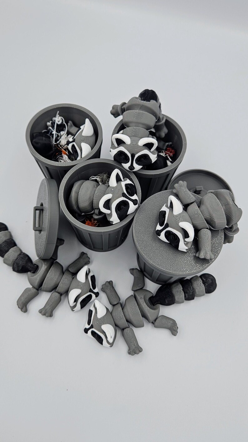 May include: A collection of gray 3D-printed raccoon figurines, some inside miniature trash cans and others disassembled. The raccoons have black and white markings. The trash cans are gray with lids and handles.