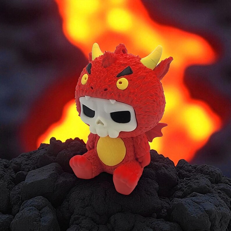 Puede incluir: Un juguete de peluche rojo con disfraz de diablo, cuernos amarillos y m&aacute;scara de calavera. Tiene un c&iacute;rculo amarillo en el pecho y est&aacute; sentado sobre una superficie oscura y texturizada, con un fondo de fuego.