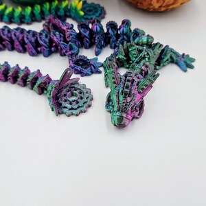 May include: A set of iridescent purple, green, and blue dragon figurines. The dragon figurines are made of plastic and have a detailed, textured surface. The figurines are arranged in a line on a white surface.