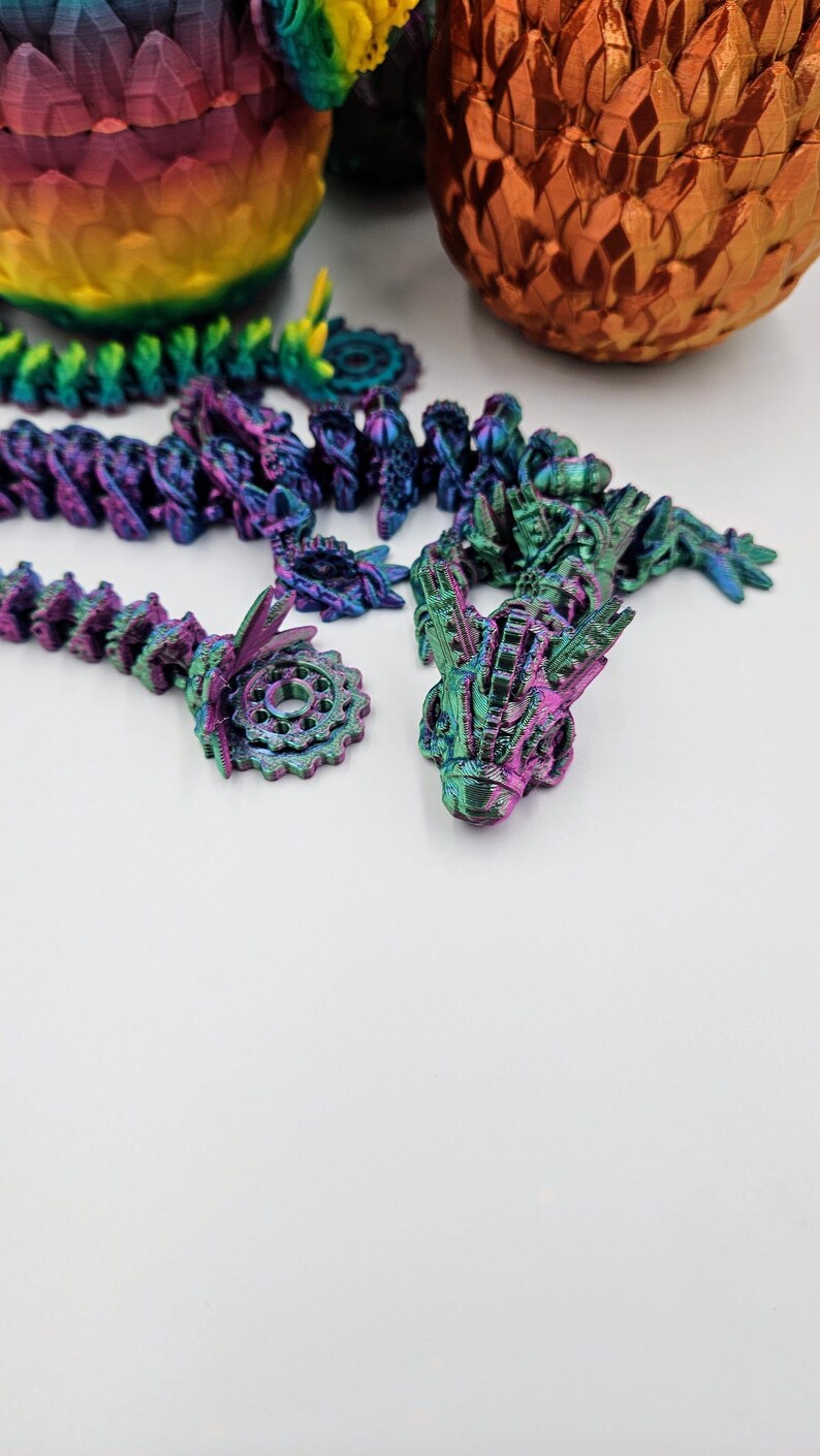 May include: A set of iridescent purple, green, and blue dragon figurines. The dragon figurines are made of plastic and have a detailed, textured surface. The figurines are arranged in a line on a white surface.