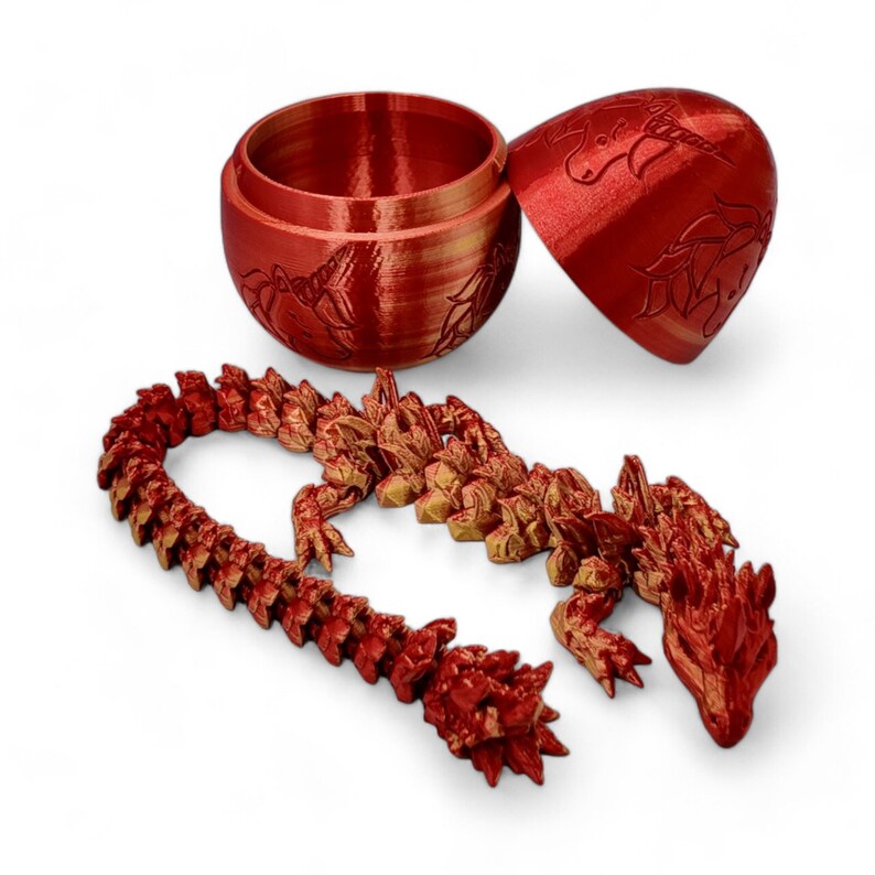 May include: A red and gold dragon figurine with a segmented body, lying on a white surface. The dragon is in a curled position with its head resting on its tail. The dragon is made of a 3D printed material and has a detailed design.