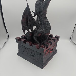 May include: A black dragon sculpture with red accents, perched atop a castle-like box. The dragon has open jaws and detailed wings. The box is dark with a brick pattern, and the overall design has a fantasy theme.