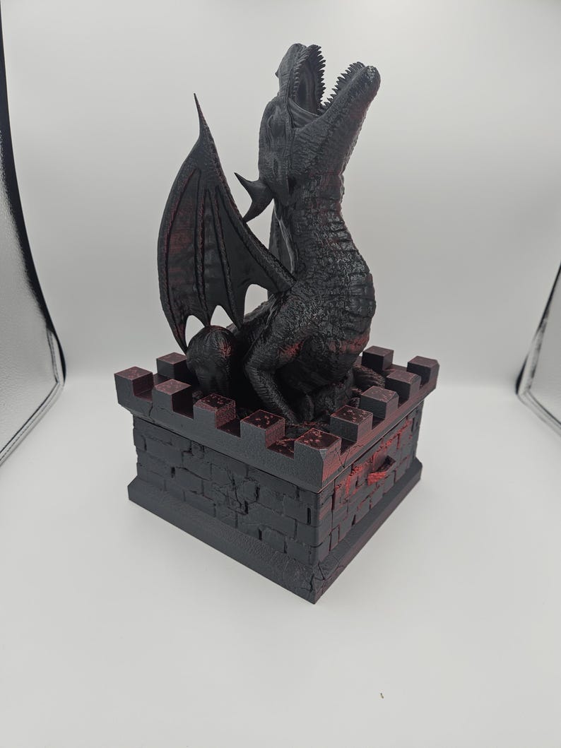 May include: A black dragon sculpture with red accents, perched atop a castle-like box. The dragon has open jaws and detailed wings. The box is dark with a brick pattern, and the overall design has a fantasy theme.