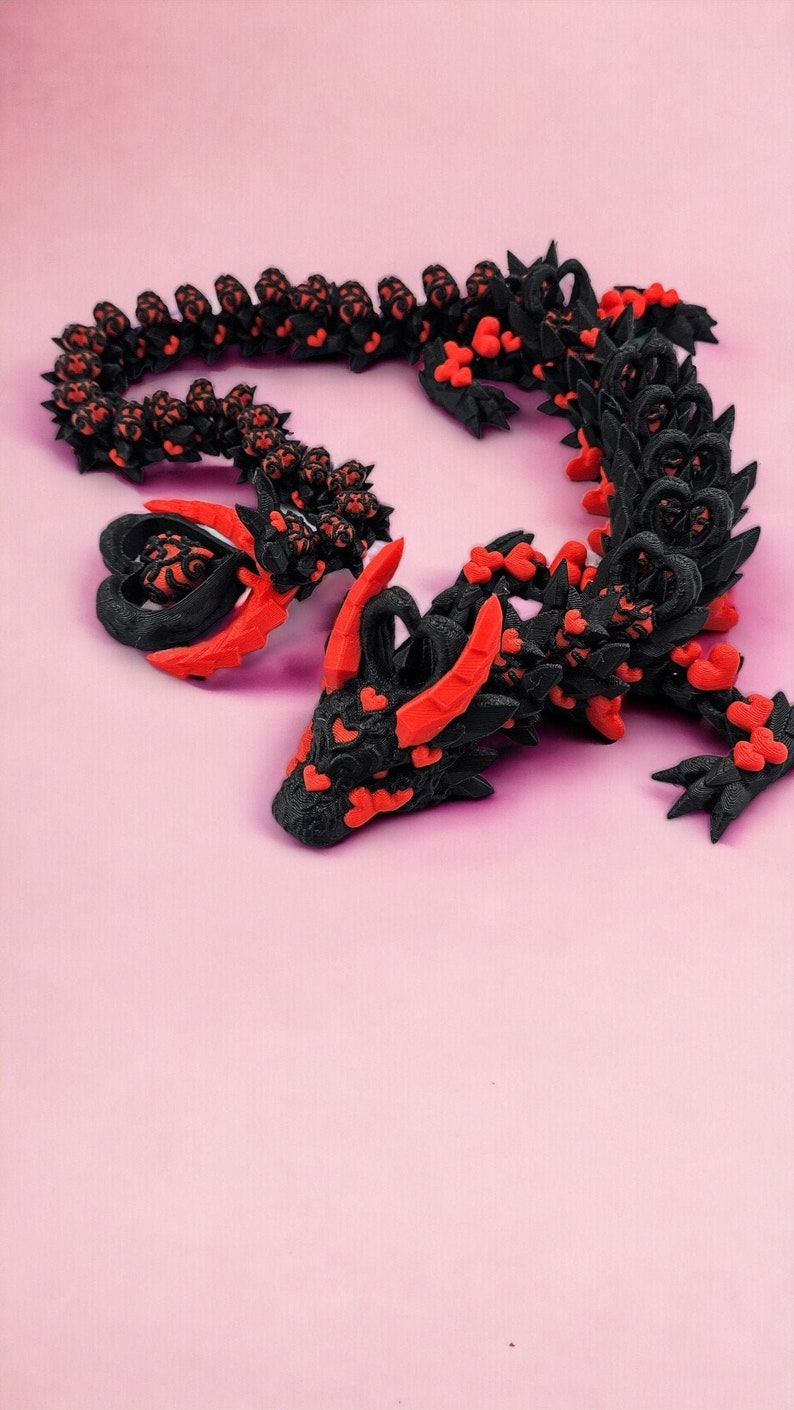 Op de afbeelding: Een zwart en rood 3D-geprint drakenbeeldje met een hartvormig ontwerp. De draak is opgerold in een cirkel met zijn kop op zijn staart.
