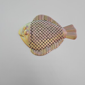 May include: A decorative fish sculpture with a textured, scale-like body. The fish features a yellow head, a body with a grid-like pattern, and a tail and fins in shades of gold and pink. The sculpture is likely made of wood or a similar material.