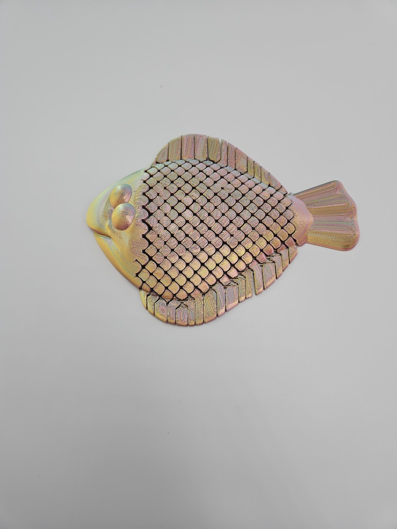 May include: A decorative fish sculpture with a textured, scale-like body. The fish features a yellow head, a body with a grid-like pattern, and a tail and fins in shades of gold and pink. The sculpture is likely made of wood or a similar material.