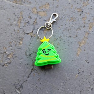 Pode incluir: Um chaveiro de &aacute;rvore de Natal verde com uma cara piscando e uma estrela amarela no topo. O chaveiro tem um anel de metal prateado e um fecho de metal prateado.