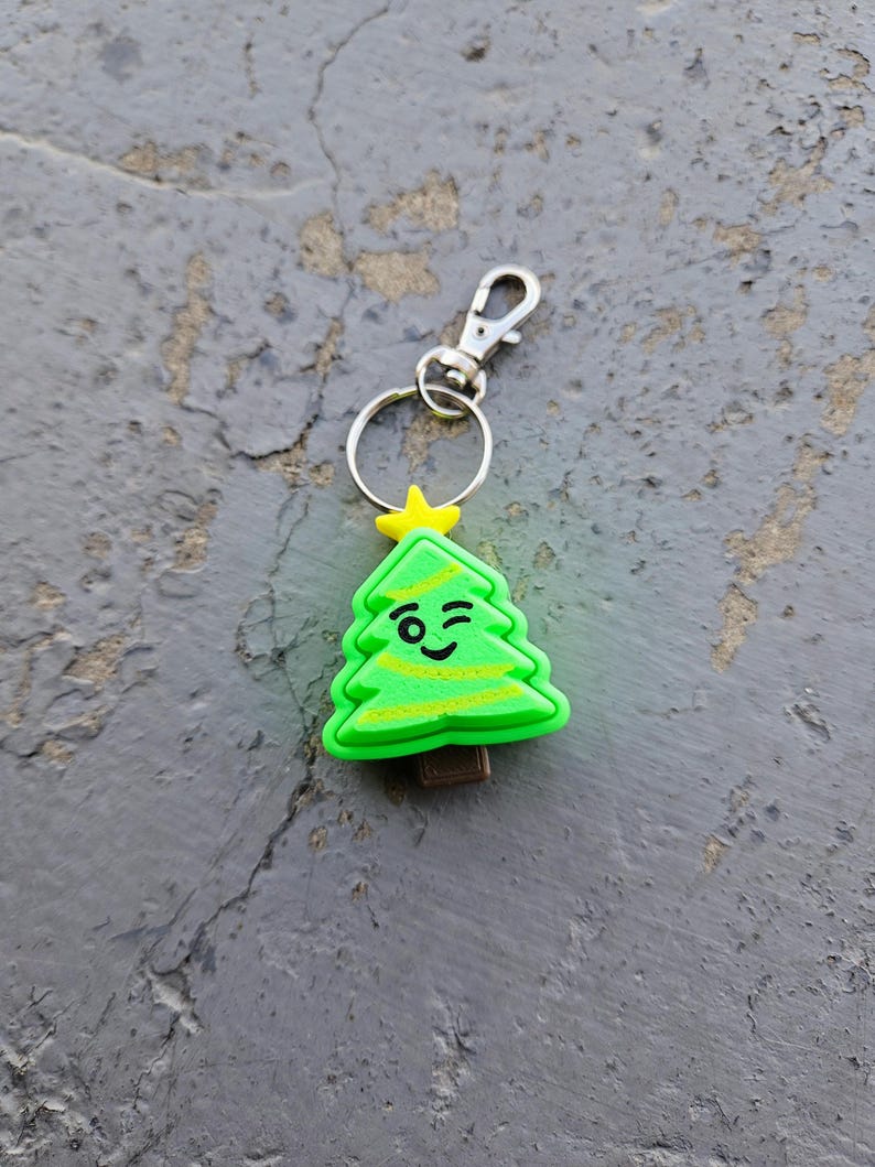 Pode incluir: Um chaveiro de &aacute;rvore de Natal verde com uma cara piscando e uma estrela amarela no topo. O chaveiro tem um anel de metal prateado e um fecho de metal prateado.
