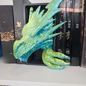 May include: A teal and gold dragon head bookend. The dragon's head and clawed foot are visible, with the head positioned between books. The bookend is made of a textured material and has a detailed design. The dragon's head is facing left.