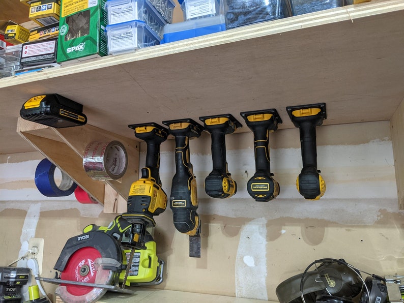 May include: A set of five yellow and black cordless power tools hanging on a wooden shelf. The tools are mounted on black brackets and are all facing the same direction. The tools are all in a row and are evenly spaced apart. There is a yellow and black battery on the shelf above the tools.