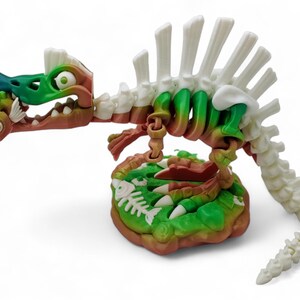 May include: A green, white, and brown plastic dinosaur skeleton toy with a fish in its mouth. The dinosaur is standing on a green and brown base with fish bones.