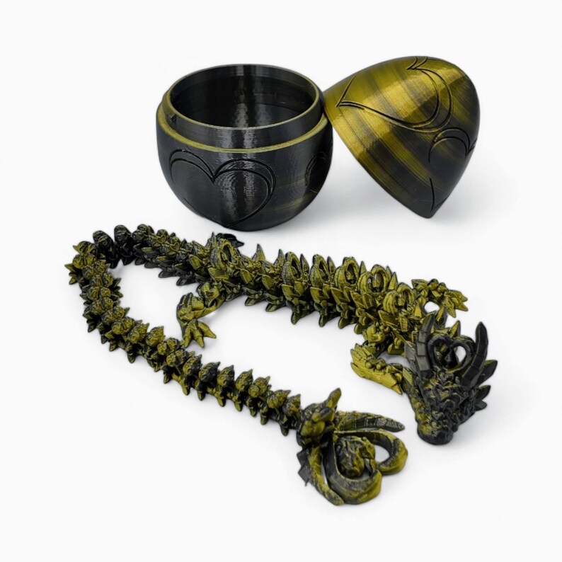 May include: A black and gold dragon figurine with a segmented body, curled into a circle. The dragon is sitting on a white surface. The dragon is part of a set that includes a black and gold egg-shaped box.