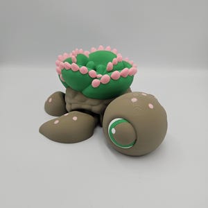 Pode incluir: Estatueta de tartaruga artesanal com carapa&ccedil;a em forma de cacto. A carapa&ccedil;a &eacute; verde com detalhes rosa, o corpo &eacute; castanho e tem um olho grande com &iacute;ris branca e verde. Um item decorativo &uacute;nico.