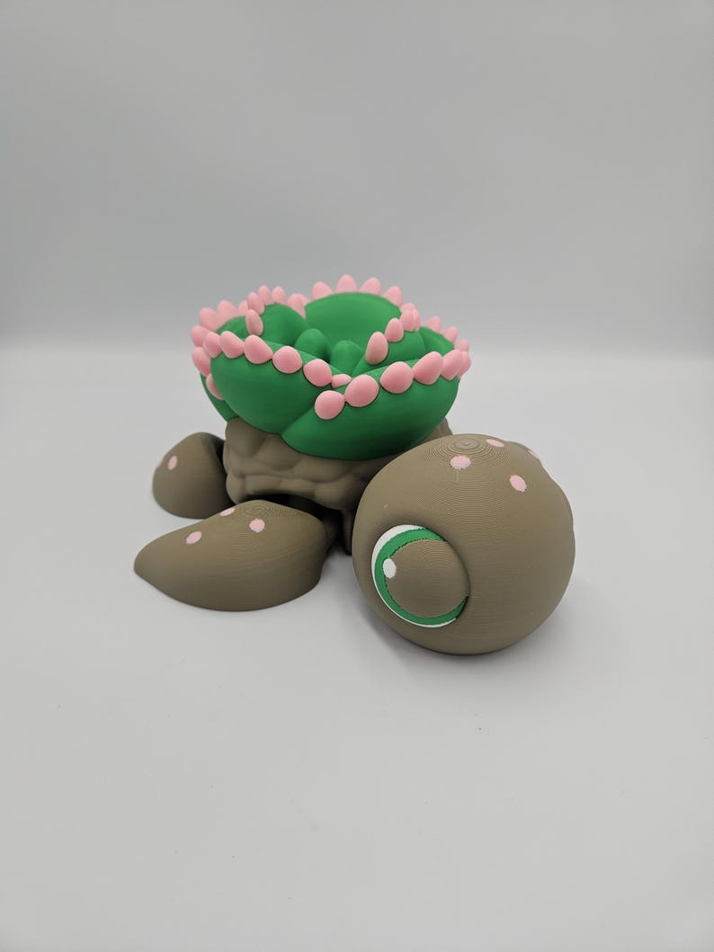 Pode incluir: Estatueta de tartaruga artesanal com carapa&ccedil;a em forma de cacto. A carapa&ccedil;a &eacute; verde com detalhes rosa, o corpo &eacute; castanho e tem um olho grande com &iacute;ris branca e verde. Um item decorativo &uacute;nico.