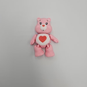 May include: A pink Care Bear figurine with a red heart on its belly. The bear has a white belly, small blue eyes, and a white nose. Its arms and legs are articulated, and it stands on a flat surface.