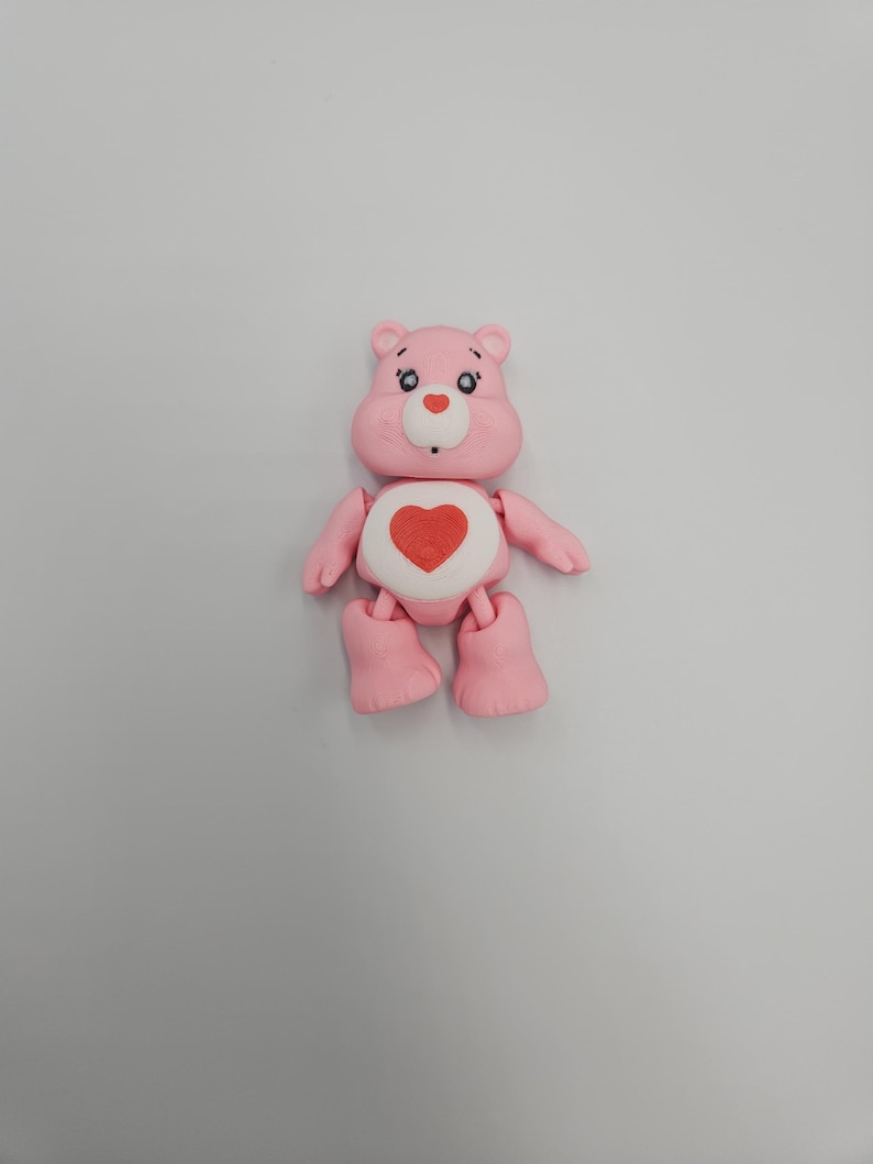 May include: A pink Care Bear figurine with a red heart on its belly. The bear has a white belly, small blue eyes, and a white nose. Its arms and legs are articulated, and it stands on a flat surface.