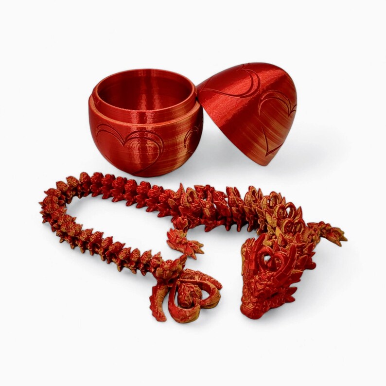 May include: A red and gold dragon figurine with a curled tail and a detailed head. The dragon is laying on a white surface. There is a red and gold egg-shaped box with a heart design on the side. The box is open and the lid is laying next to the dragon.