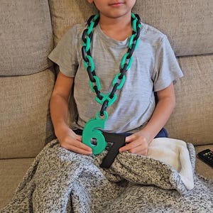 May include: A child wearing a green and black chain necklace with a large green number 6 pendant and a black number 7. The necklace is made of large, interlocking links. The child is sitting on a couch.