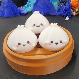 May include: Three white, cartoon-style dumplings with smiling faces and pink cheeks sit in a brown, wooden-looking steamer basket. The dumplings have black eyes and small, black mouths. The steamer basket has a round shape.