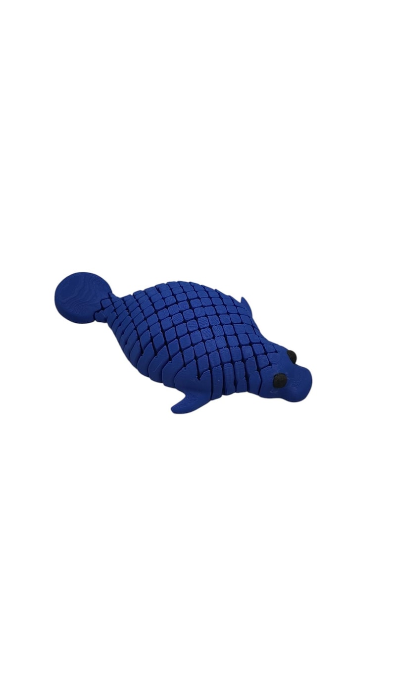 Puede incluir: Una escultura azul estilizada de una foca. La foca tiene un patr&oacute;n texturizado, similar a escamas, en su cuerpo, con peque&ntilde;os ojos negros. La escultura est&aacute; sobre un fondo blanco liso.