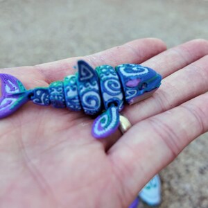 May include: A small, blue and purple, segmented shark toy with a white and blue patterned design. The toy is made of plastic and has a detailed design.