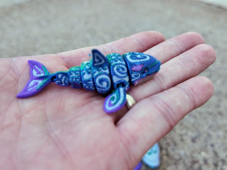 May include: A small, blue and purple, segmented shark toy with a white and blue patterned design. The toy is made of plastic and has a detailed design.