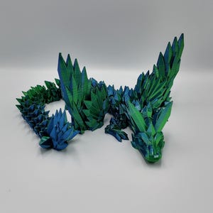 May include: A 3D printed dragon sculpture made of green and blue plastic. The dragon is posed in a curled position with its wings spread out.