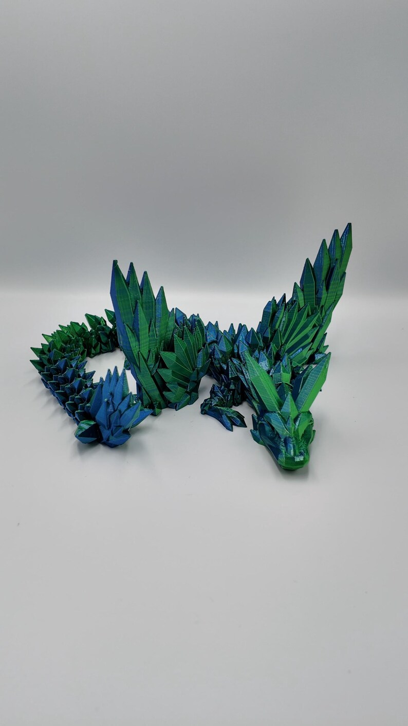 May include: A 3D printed dragon sculpture made of green and blue plastic. The dragon is posed in a curled position with its wings spread out.