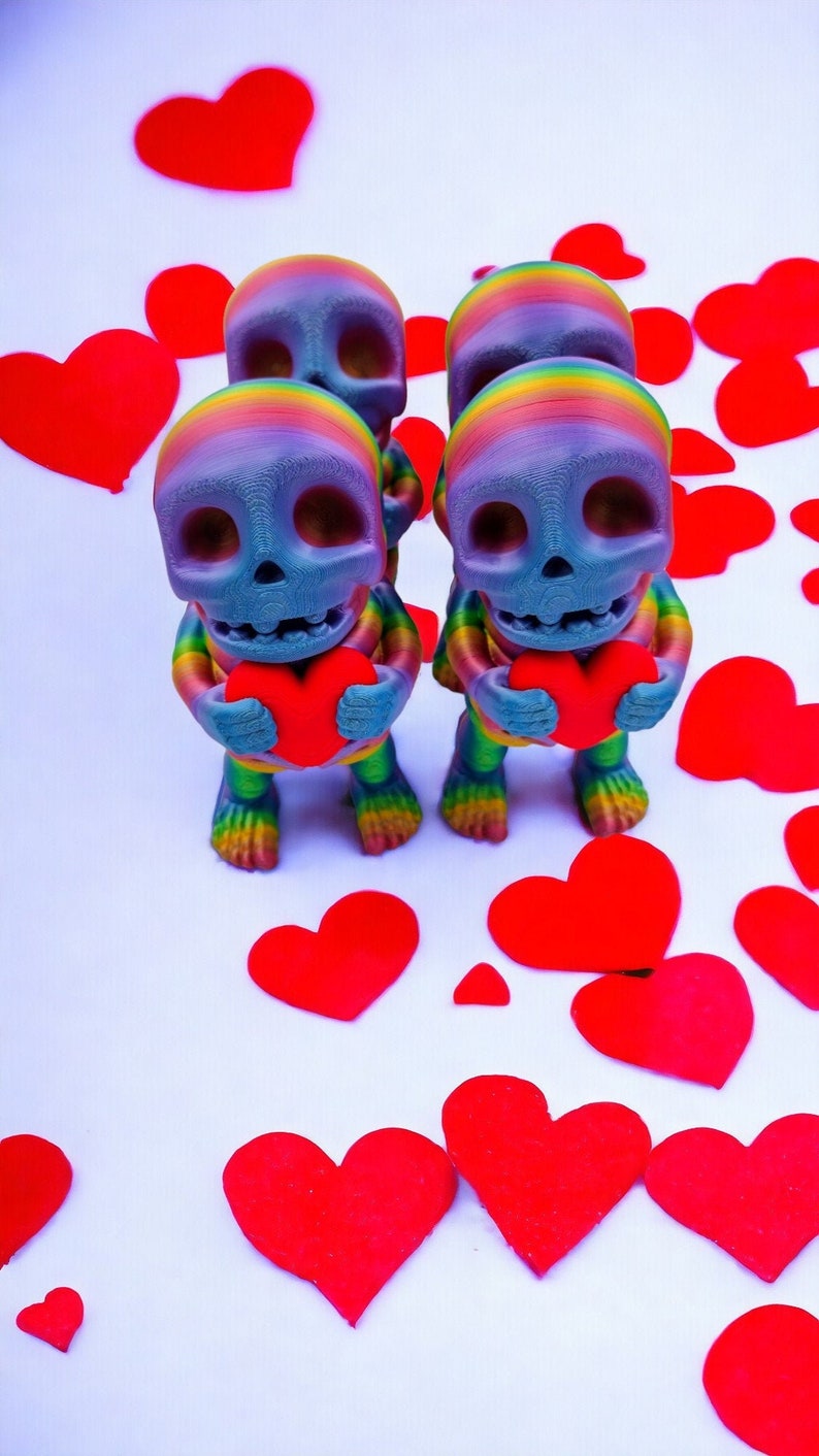 May include: Three rainbow-coloured skull figurines holding red hearts. The figurines are standing on a white surface surrounded by red paper hearts.
