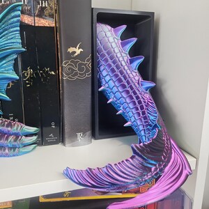 May include: A mermaid tail-shaped bookend in shades of purple and blue. The bookend has a textured, scaled pattern and sharp dorsal fins. It is placed between books on a white shelf, adding a fantasy touch to home decor.