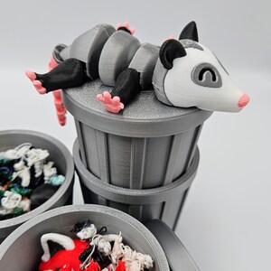 May include: A 3D-printed gray trash can set with a possum figurine. The possum has a white face, black ears, and pink feet. The trash cans are filled with colorful, tangled yarn.