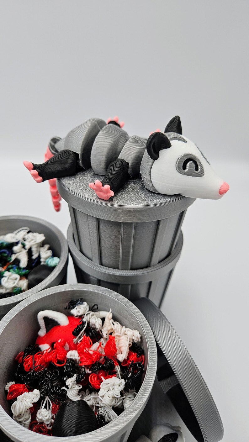 May include: A 3D-printed gray trash can set with a possum figurine. The possum has a white face, black ears, and pink feet. The trash cans are filled with colorful, tangled yarn.