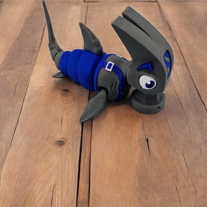 May include: A gray and blue 3D printed toy shark with a cartoonish expression. The shark is lying on a wooden surface.
