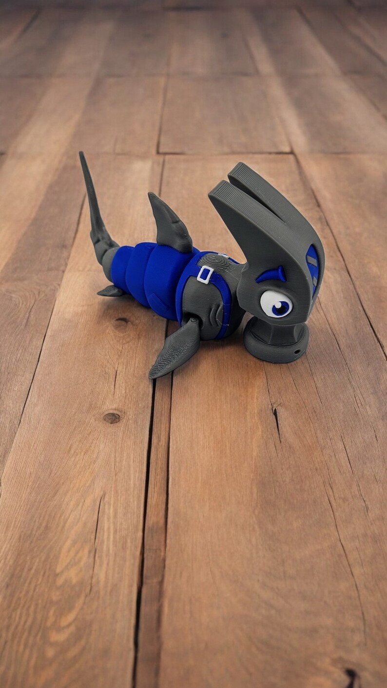 May include: A gray and blue 3D printed toy shark with a cartoonish expression. The shark is lying on a wooden surface.