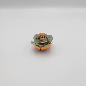 May include: A single, artificial rose with a multi-colored design. The petals transition from a light teal to a warm orange-brown in the center, with yellow and orange at the base. The rose is centered on a white background.