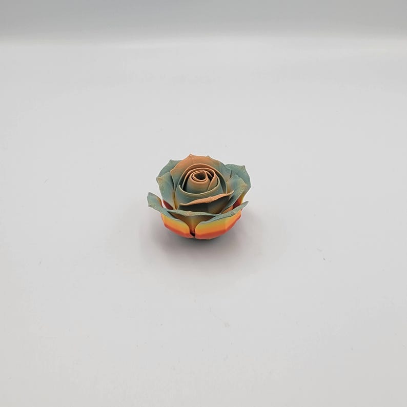 May include: A single, artificial rose with a multi-colored design. The petals transition from a light teal to a warm orange-brown in the center, with yellow and orange at the base. The rose is centered on a white background.