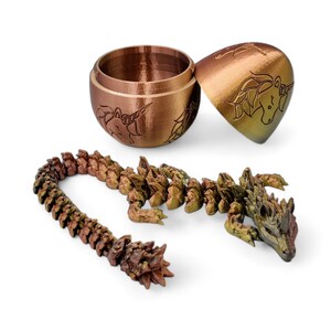 May include: A 3D printed dragon figurine with a segmented tail, a detailed head, and a metallic bronze and gold color scheme. The dragon is curled up and appears to be made of multiple pieces. The dragon is next to a closed, round, metallic bronze and gold egg-shaped container with a unicorn design.