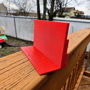 May include: A bright red plastic stand with a stepped design. The stand is made of 3D printed plastic and is sitting on a wooden surface.