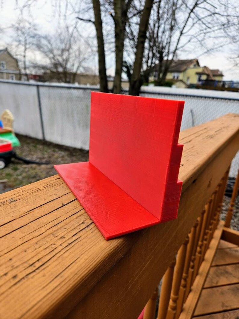 May include: A bright red plastic stand with a stepped design. The stand is made of 3D printed plastic and is sitting on a wooden surface.