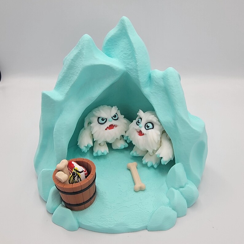 May include: A whimsical diorama featuring two white, furry creatures with red mouths and blue eyes, nestled inside a light blue ice cave. A small wooden barrel and a bone are also present, creating a playful scene.