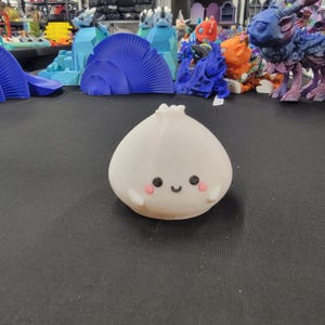 Peut inclure: Figurine en forme d'ail blanc de style cartoon, avec un visage souriant, des joues roses et de petits bras. La figurine est plac&eacute;e devant un fond de diverses cr&eacute;atures et structures fantastiques imprim&eacute;es en 3D.