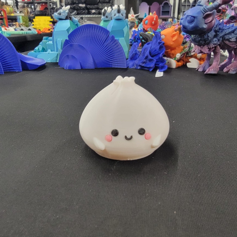 Peut inclure: Figurine en forme d'ail blanc de style cartoon, avec un visage souriant, des joues roses et de petits bras. La figurine est plac&eacute;e devant un fond de diverses cr&eacute;atures et structures fantastiques imprim&eacute;es en 3D.