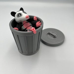 Op de afbeelding: Een grijze 3D-geprinte vuilnisbak met bijpassend deksel, naast een cartoon opossum beeldje. De opossum heeft een wit gezicht, roze neus en poten, en zwarte oren en ledematen. De opossum zit in de vuilnisbak.