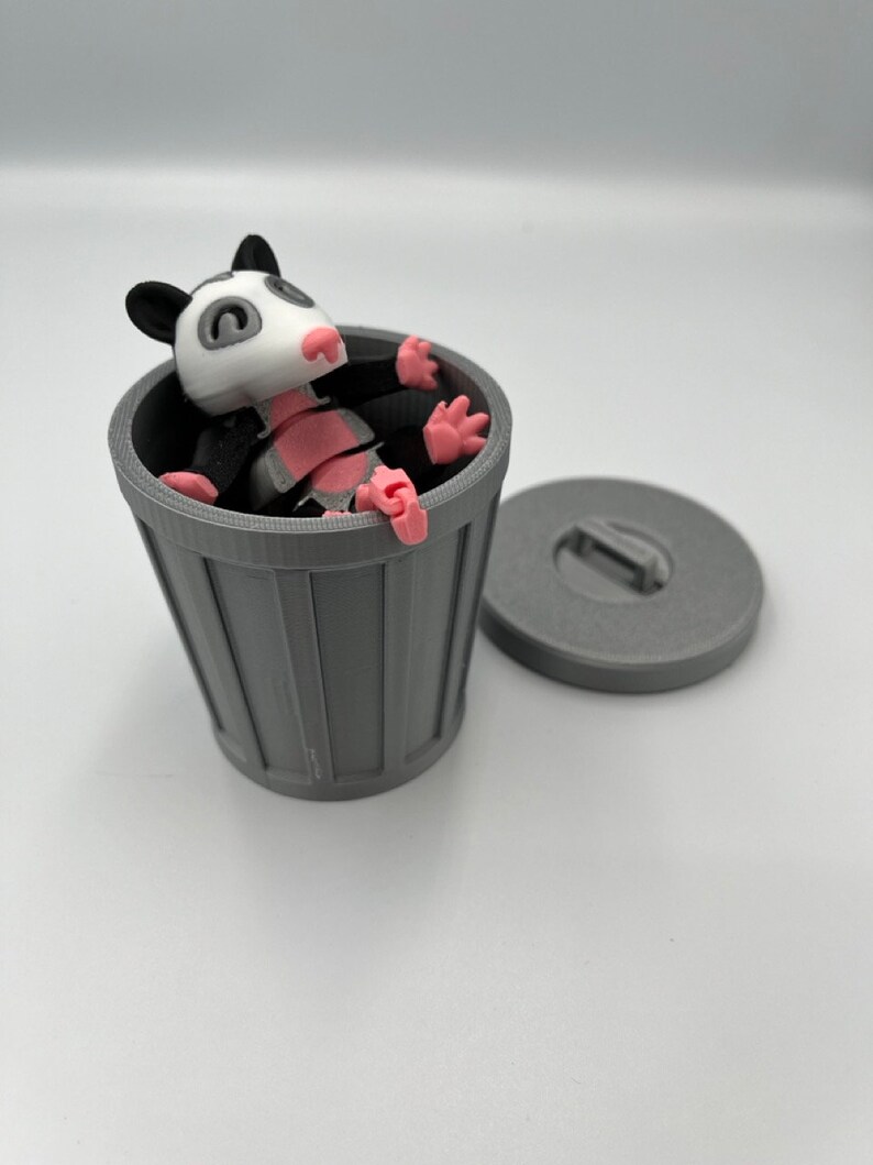 Op de afbeelding: Een grijze 3D-geprinte vuilnisbak met bijpassend deksel, naast een cartoon opossum beeldje. De opossum heeft een wit gezicht, roze neus en poten, en zwarte oren en ledematen. De opossum zit in de vuilnisbak.