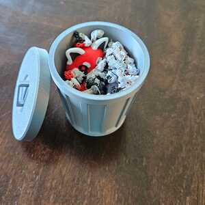 May include: A light blue 3D-printed miniature trash can with a lid, filled with small, colorful objects. The objects include red, black, and white shapes. The lid is slightly ajar, revealing the contents.