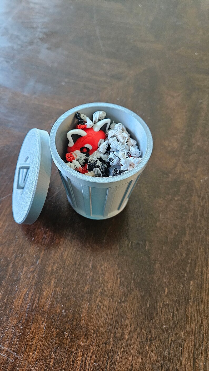 May include: A light blue 3D-printed miniature trash can with a lid, filled with small, colorful objects. The objects include red, black, and white shapes. The lid is slightly ajar, revealing the contents.
