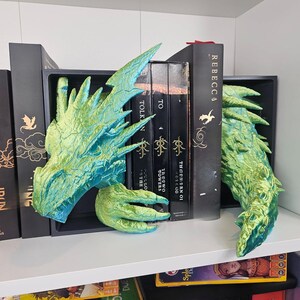 May include: Dragon-themed bookends in shades of green and blue, holding up a collection of books. The bookends are designed to resemble a dragon's head and tail. The books have gold lettering and include the title "REBECCA".
