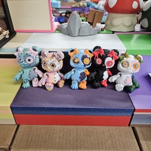 May include: Six handmade teddy bear figurines with distinct designs. The bears are in various colours such as blue, pink, orange, black, and white. They have button eyes and decorative details, presented on a purple and red surface.