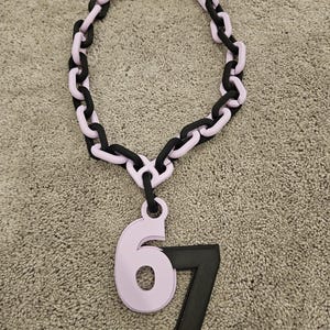 May include: A chunky necklace featuring a chain design with alternating black and light purple links. A light purple pendant with the number 6 and a black number 7 hangs from the chain.